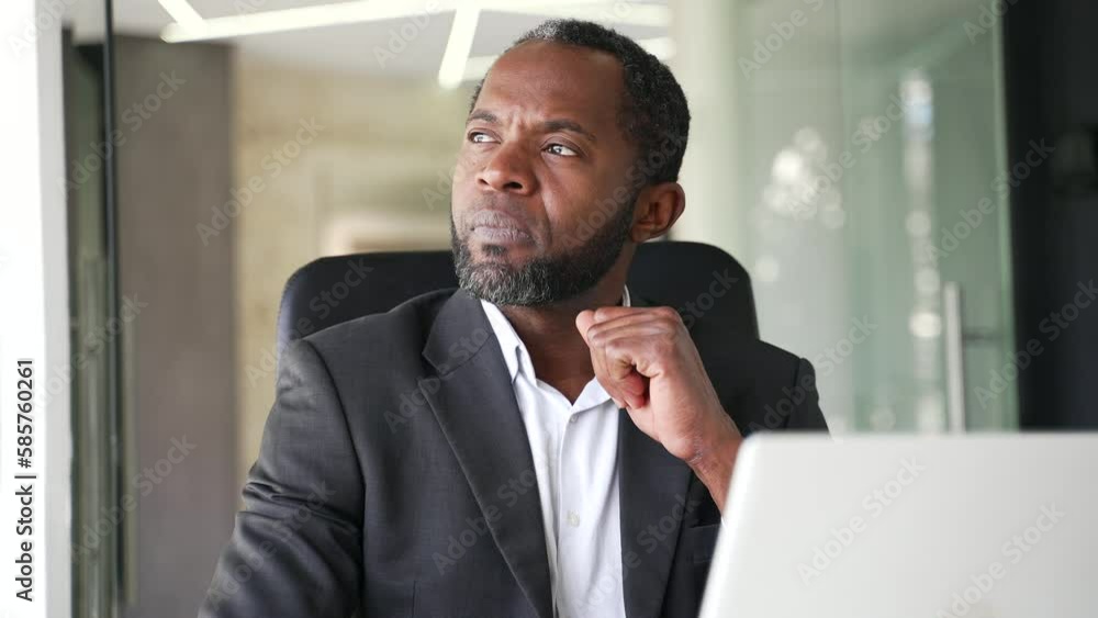 Close up of a thoughtful mature african american man in a formal suit sitting at a workplace in a office using a laptop. Concentrated middle aged businessman is thinking about a serious business deal Close up of a thoughtful mature african american man in a formal suit sitting at a workplace in a office using a laptop. Concentrated middle aged businessman is thinking about a serious business deal