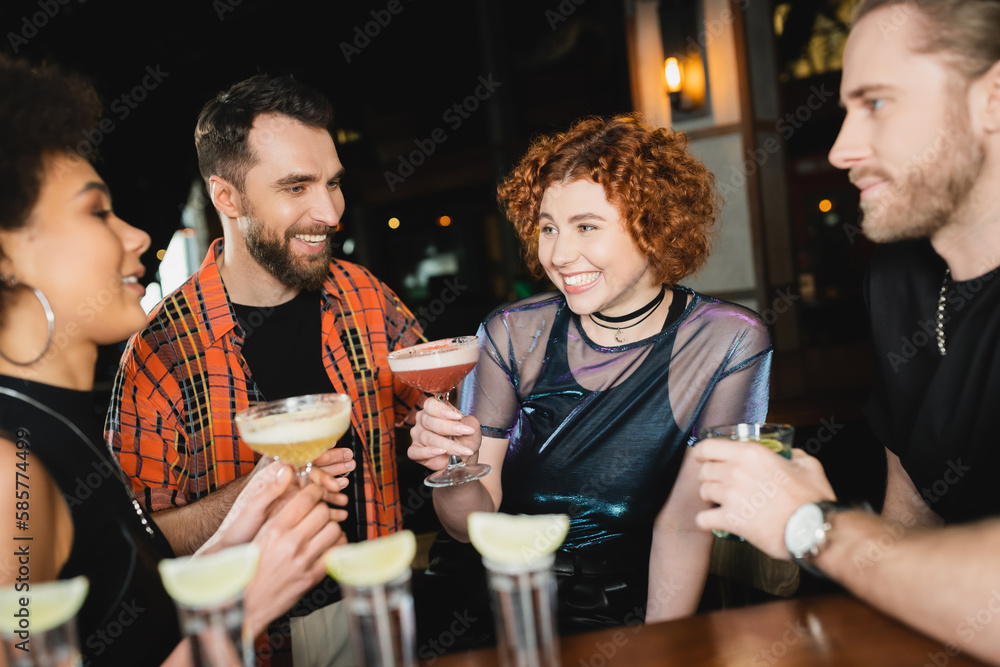 Cheerful woman holding cocktail near multiethnic friends and tequila shots in bar.