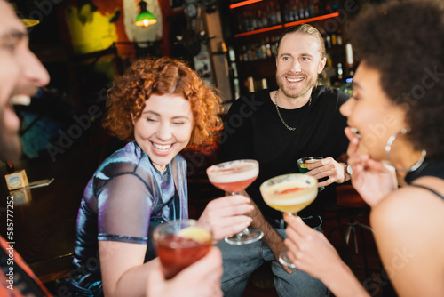 Positive man holding blue lagoon cocktail near interracial friends talking in bar.