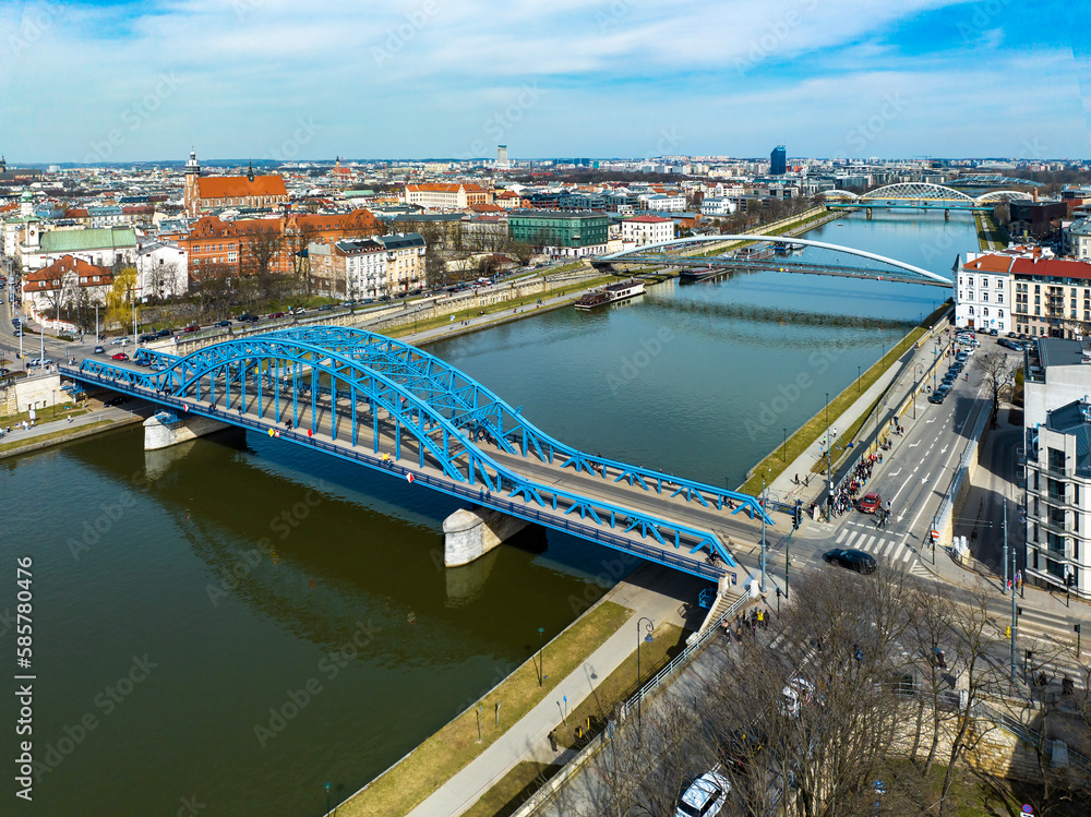 Obraz premium Bridges on Vistula River in Krakow, Poland. Aerial view. Boulevards with waking people. Blue tied-arc bridge in front. Footbridge for pedestrians and cyclist. Far view of new double railway bridge