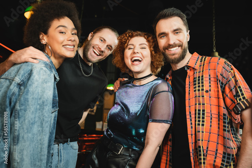 Positive multiethnic friends hugging and looking at camera in bar at night.