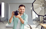 Portrait of a young happy smiling photographer in casual clothes holding a camera in his hands going take a shoot standing in professionally production studio with light equipment.