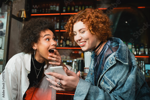 Excited redhead woman using smartphone near african american friend in bar.