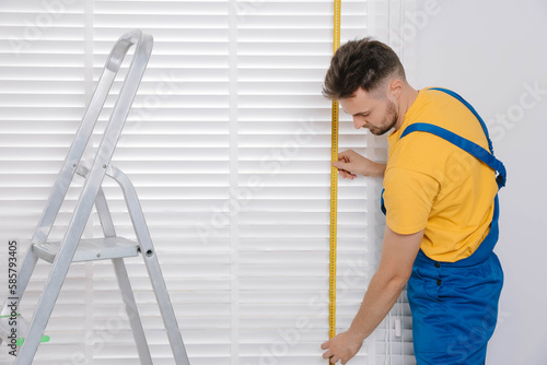 Worker in uniform using measuring tape while installing horizontal window blinds near stepladder indoors