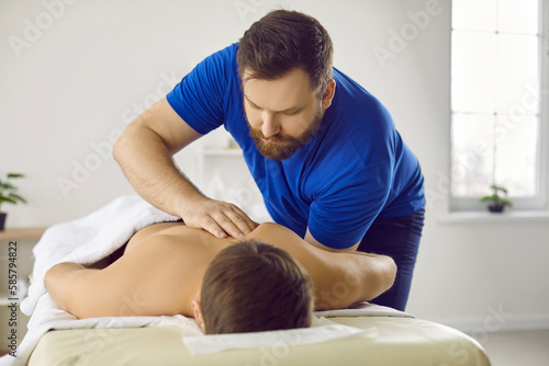 Male masseur do manual massage to client lying on table in studio or clinic. Attentive professional physiotherapist work with customer back relieve pain in stiff muscles. Physiotherapy.