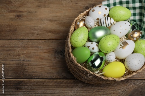 Many beautifully decorated Easter eggs in wicker basket on wooden table, above view. Space for text