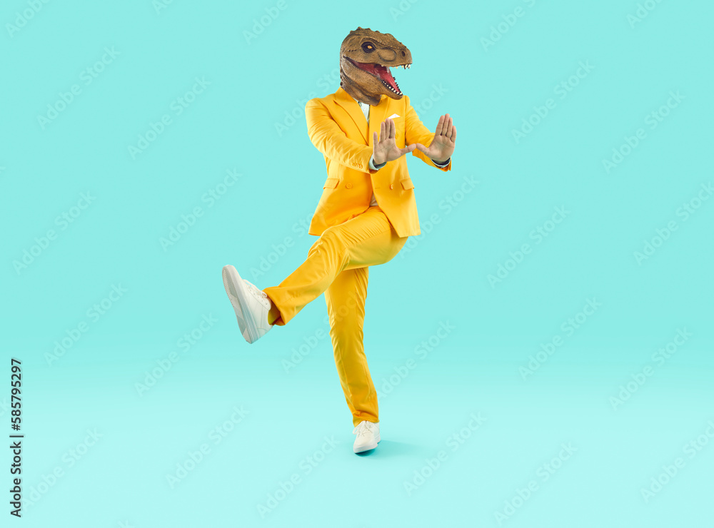Funny man in a dinosaur costume dancing in the studio. Happy guy in a bright yellow suit and funny dinosaur mask dancing on a turquoise blue colour background. Having fun at a party concept