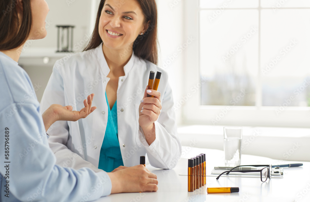 Homeopathic doctor suggesting young woman to choose scent of essential oil in medical clinic. Doctor holding herbal essential oil flasks while talking to female patient. Nature aromatherapy concept Homeopathic doctor suggesting young woman to choose scent of essential oil in medical clinic. Doctor holding herbal essential oil flasks while talking to female patient. Nature aromatherapy concept