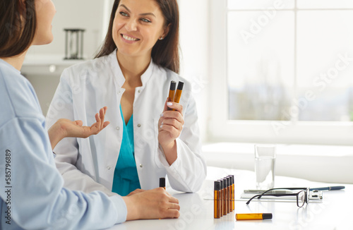 Homeopathic doctor suggesting young woman to choose scent of essential oil in medical clinic. Doctor holding herbal essential oil flasks while talking to female patient. Nature aromatherapy concept