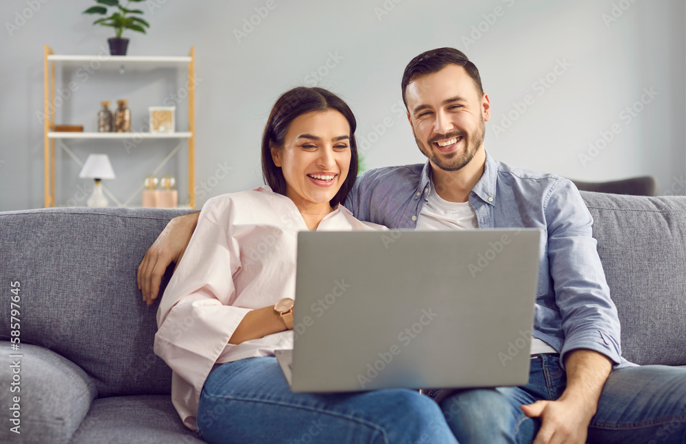 Happy smiling young couple are sitting together on cozy sofa at home in living room with laptop in their hands, looking at the monitor screen. Feeling of joy, emotional excitement. Celebrate success. Happy smiling young couple are sitting together on cozy sofa at home in living room with laptop in their hands, looking at the monitor screen. Feeling of joy, emotional excitement. Celebrate success.