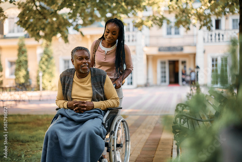 Happy black woman pushes her senior father in wheelchair while visiting his at nursing home.