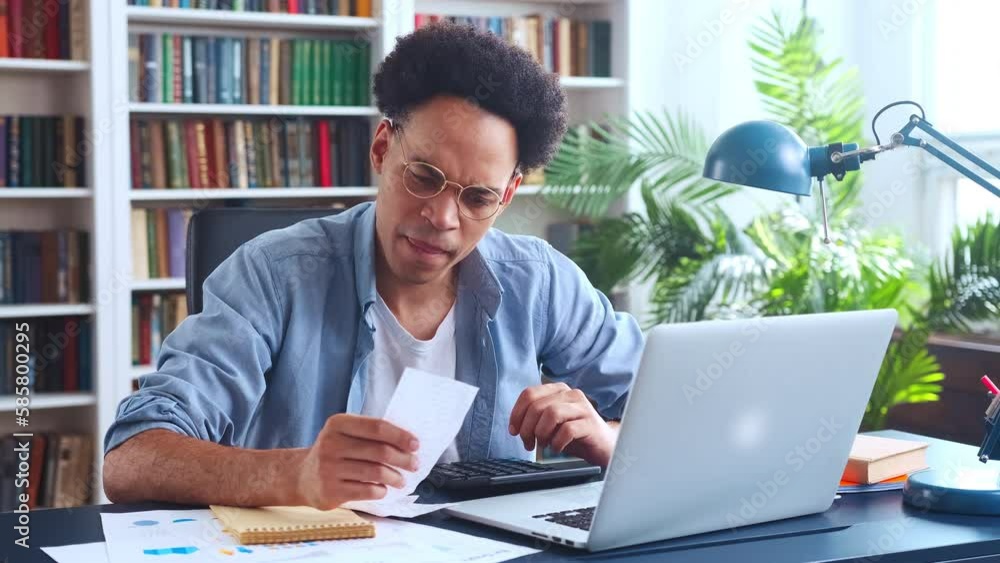 Young focused smart African American man auditor enters data on financial expenses into laptop by retyping amounts from cashier checks into CPM system sits at desk in home office. Accounting, finance Young focused smart African American man auditor enters data on financial expenses into laptop by retyping amounts from cashier checks into CPM system sits at desk in home office. Accounting, finance