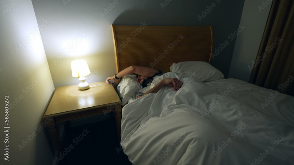 Obraz premium Man laying down in bed to rest after long day. Person going to sleep going under bedsheets resting turning bedside lamp off