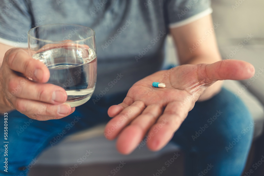 Elderly man taking pills for depression sitting on couch. Old upset patient swallowing pill with glass of water.
