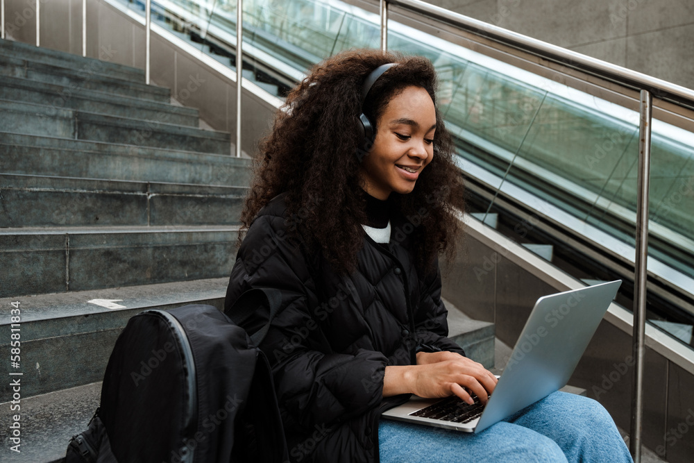 Fototapeta premium Young african girl working on laptop while sitting on stairs