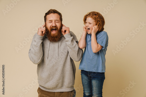 Father and son covering their ears with fingers and screaming while standing isolated over beige background