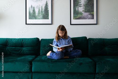 portrait of a schoolgirl in blue shirt sitting on sofa with book at home