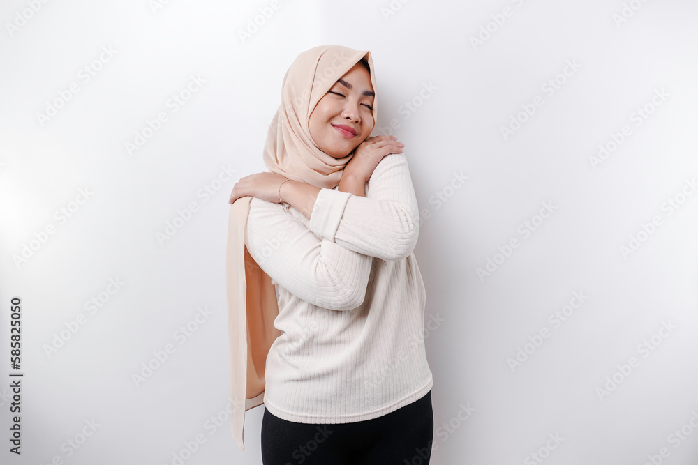 Fototapeta premium Young beautiful Asian Muslim woman wearing a headscarf over white background hugging herself happy and positive, smiling confident. Self-love and self-care