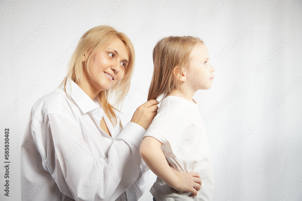 Close portrait of blonde mother and daughter where mom braids hair and makes ponytail and hairstyle for her girl on white background in studio. The concept of love, friendship, caring in the family Close portrait of blonde mother and daughter where mom braids hair and makes ponytail and hairstyle for her girl on white background in studio. The concept of love, friendship, caring in the family