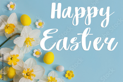 Happy Easter greeting card. Happy Easter text on stylish Easter eggs and yellow daffodils flowers flat lay on blue background. Modern Greeting card. Handwritten sign © sonyachny