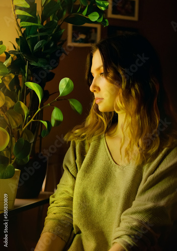Portrait of a beautiful young blonde woman who is sitting at home in the evening near indoor green plants. Home gardening and love of plants. Ecology.