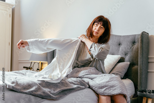 Unhappy 55s lady feel disturbed frustrated suffer from insomnia concept uncomfortable bad mattress sits on the bed puts on a bathrobe.