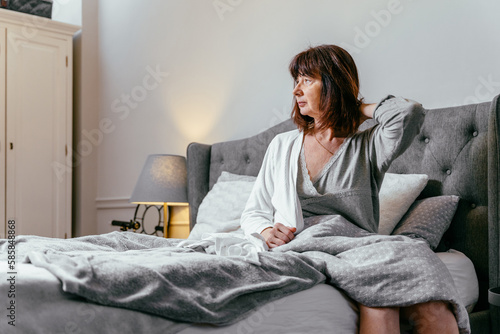 Tired middle aged woman sits on the bed puts on a bathrobe can't sleep late at morning with insomnia. Adult lady sick or sad depressed sleeping at home. photo