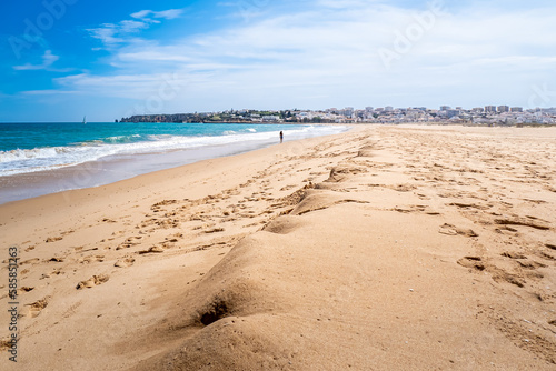 At low tide, Beach Meia Praia stretches far and wide with empty shoreline and calm waves, with an unrecognizable woman in the distance and the charming town of Lagos visible in the background.