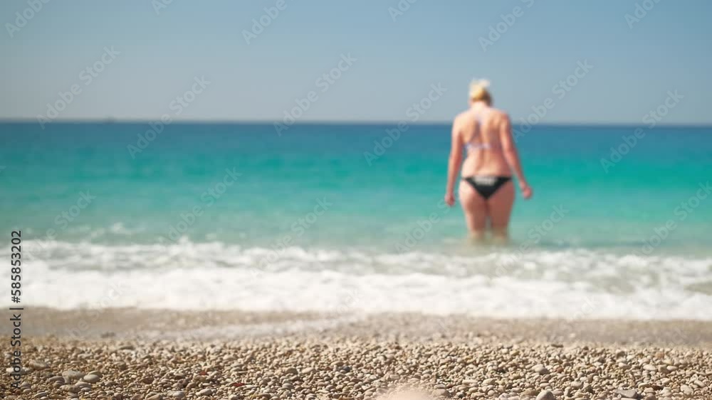 Blurred background of live video of a magnificent view of the beach on the Aegean Sea, focus on the pebbles on the beach, a beautiful adult woman is standing in the sea, a bright sunny day Blurred background of live video of a magnificent view of the beach on the Aegean Sea, focus on the pebbles on the beach, a beautiful adult woman is standing in the sea, a bright sunny day