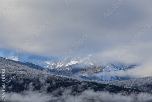 A scenic view of majestic snowy and foggy mountain with some frost on the top of the forest under a majestic blue sky