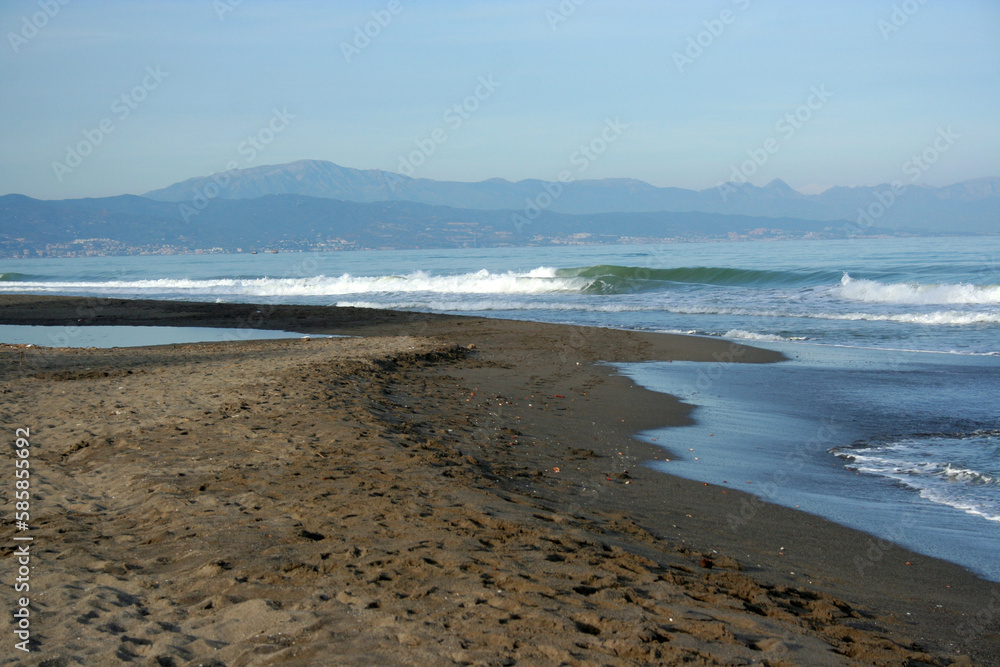 sandy beach with sand, waves skidding, mountain in the background, blue sky, end of day light