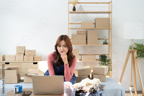 Young woman sitting and using laptop to sell products online with order packages waiting to deliver behind her. Concept of shopping online, e-commerce store, checking order. Selling products online. photo