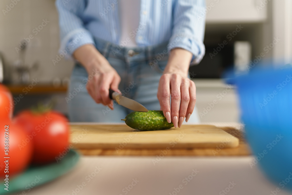 Woman cutting green cucumber on a wooden board at home. Female cooking fresh salad with vegetables. Person preparing healthy vegetarian meal Woman cutting green cucumber on a wooden board at home. Female cooking fresh salad with vegetables. Person preparing healthy vegetarian meal