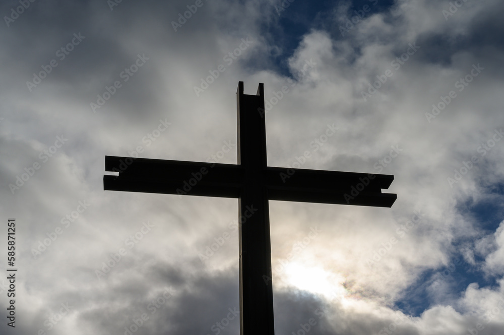Steel cross against cloudy sky with the sun breaking through and patches of blue