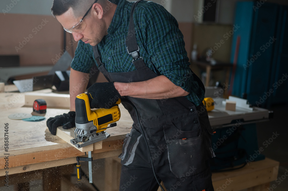 A man cuts a wooden plank with an electric jigsaw in a workshop.