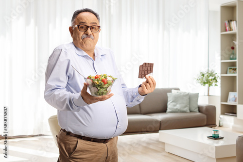Doubtful mature man holding a salad and a chocolate