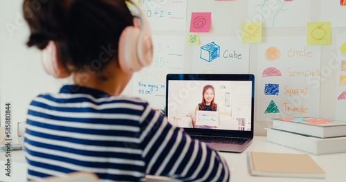 Asian toddler girl with sweater wear headphone sit front of desk with notepad use magic pen focus on writing do homework listen teacher online learning english course from laptop screen at home.