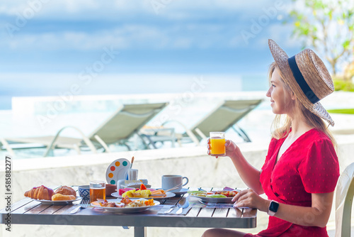 Young woman on summer vacation enjoying breakfast with tropical fruits, drinking orange juice on a luxury hotel resort terrace overlooking the sea.