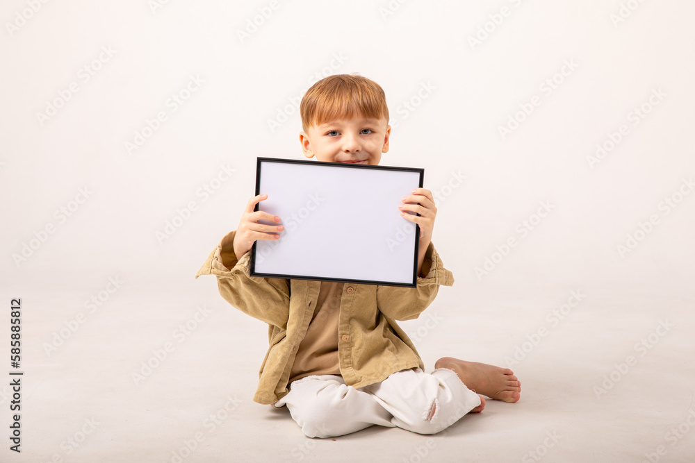 Smiling little boy holding placard on white background. Happy cute boy holding blank poster sitting on white floor. Laughing child showing empty white sign. Smiling little boy holding placard on white background. Happy cute boy holding blank poster sitting on white floor. Laughing child showing empty white sign.