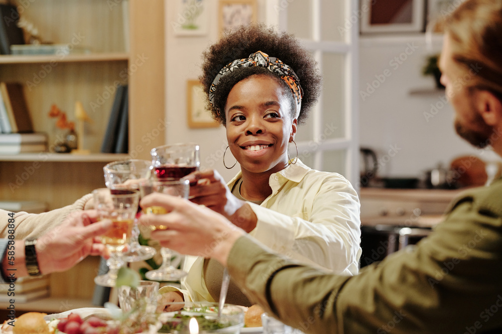 Happy young African American woman looking at her husband while clinking with wineglasses of family members during toast Happy young African American woman looking at her husband while clinking with wineglasses of family members during toast