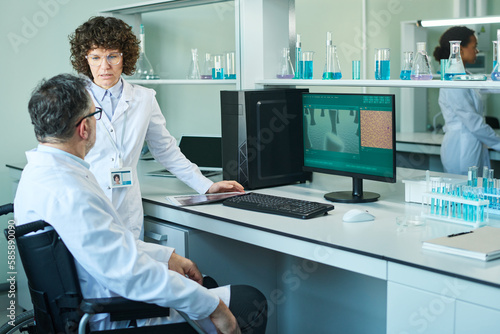 Two mature colleagues in lab coats discussing working points while woman standing in front of man with disability sitting in wheelchair