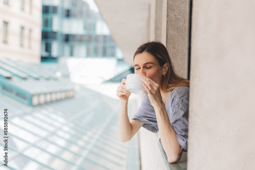 Young woman looking out the window and holding mug at home or hotel room. She drinking coffee or tea after wake up in the morning. Woman look enjoy and happy, positive mood, smell of morning coffee.