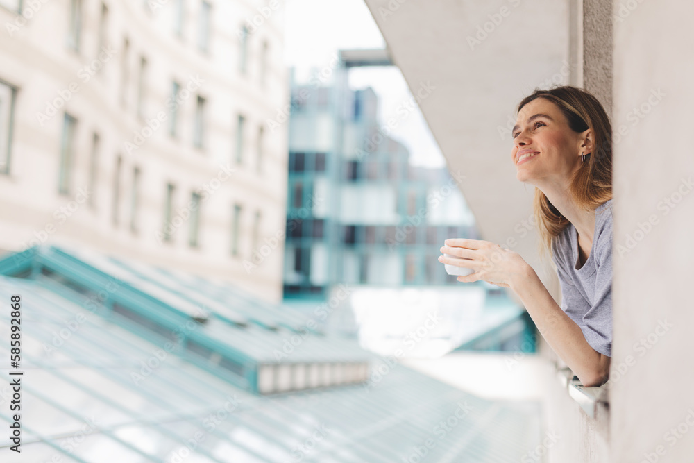 Fototapeta premium Young woman looking out the window and holding mug at home or hotel room. She drinking coffee or tea after wake up in the morning. Good morning! Woman look happy, positive mood, sitting on windowsill.