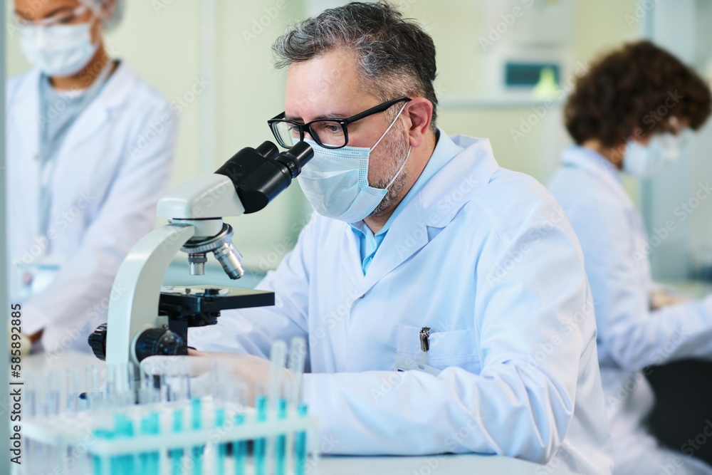 Experienced scientist in protective mask studying new virus cells in microscope while sitting by workplace in front of camera Experienced scientist in protective mask studying new virus cells in microscope while sitting by workplace in front of camera