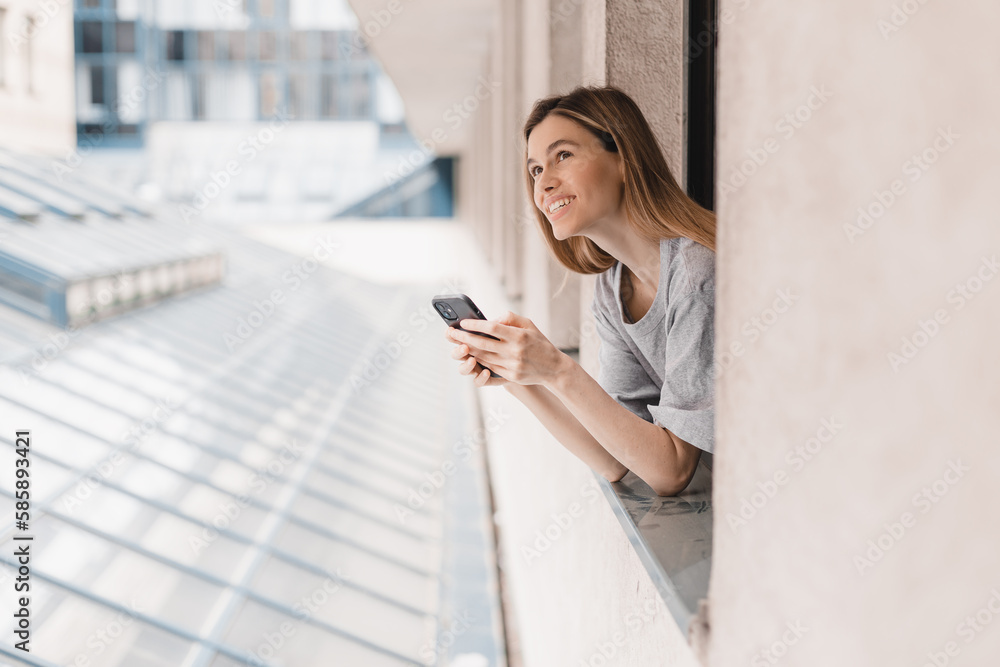 Fototapeta premium Young woman looking out the window and holding mug at home or hotel room. She drinking coffee or tea after wake up in the morning. Good morning! Woman look happy, positive mood, sitting on windowsill.