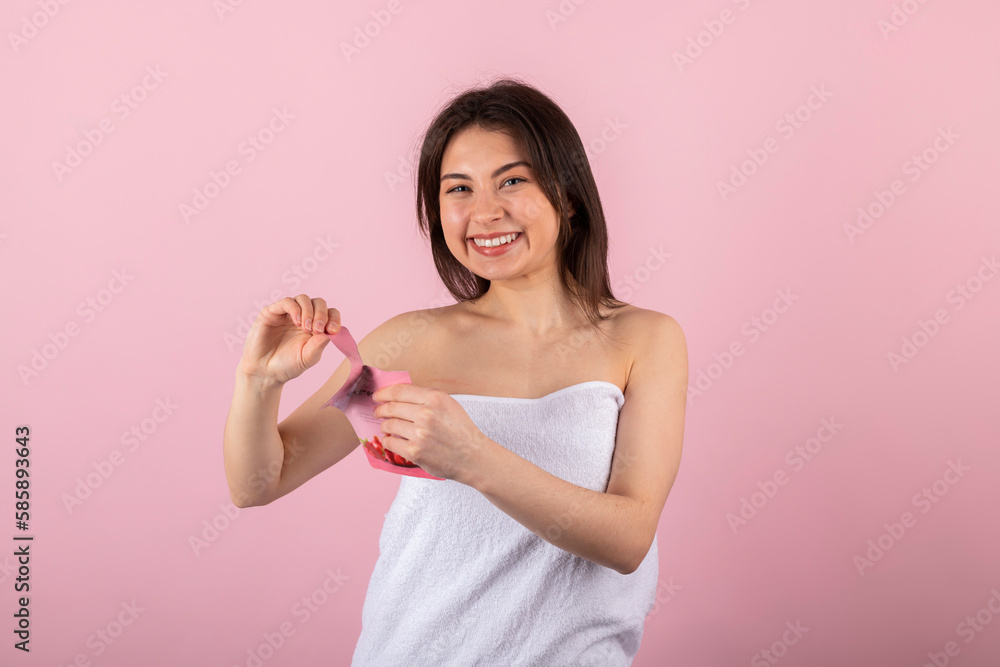 Attractive teenage girl wrapped only in a white towel happily smiling and opening her new package with a facial sheet mask or cosmetics. isolated on a pink background. Attractive teenage girl wrapped only in a white towel happily smiling and opening her new package with a facial sheet mask or cosmetics. isolated on a pink background.