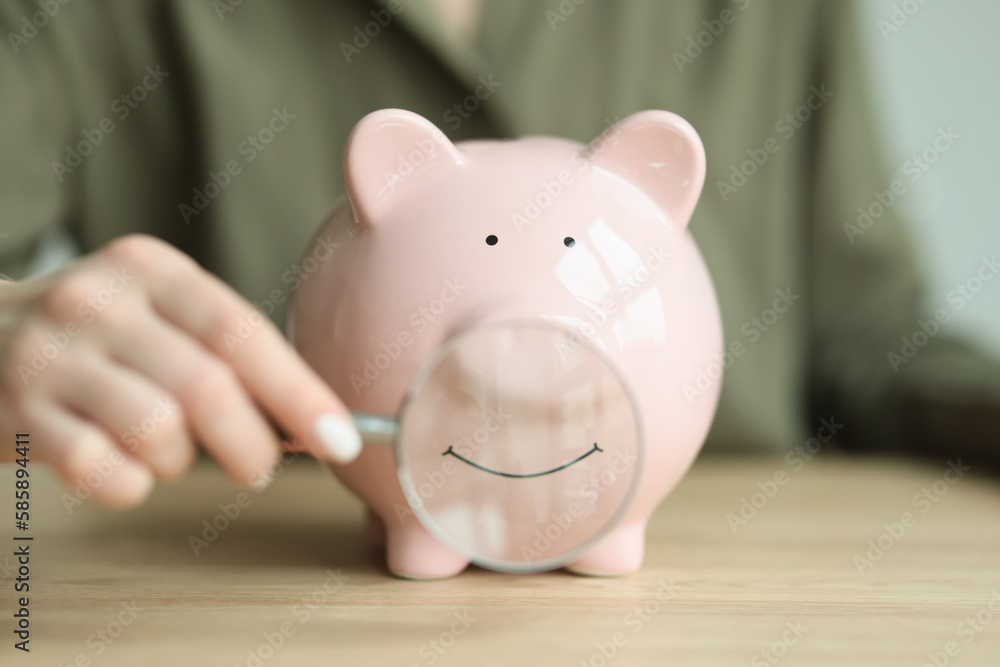 Hand of woman putting magnifier glass to mouth of piggy bank Hand of woman putting magnifier glass to mouth of piggy bank