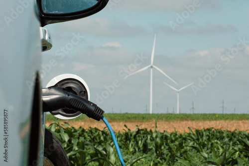 Electric car with EV plug on station with renewable form of energy produced on windfarms. Automobile stands against wind turbines on field
