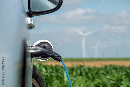 Electric car with EV plug charges from station with clean form of energy produced on windfarms. Automobile stands against blurred wind turbines on field
