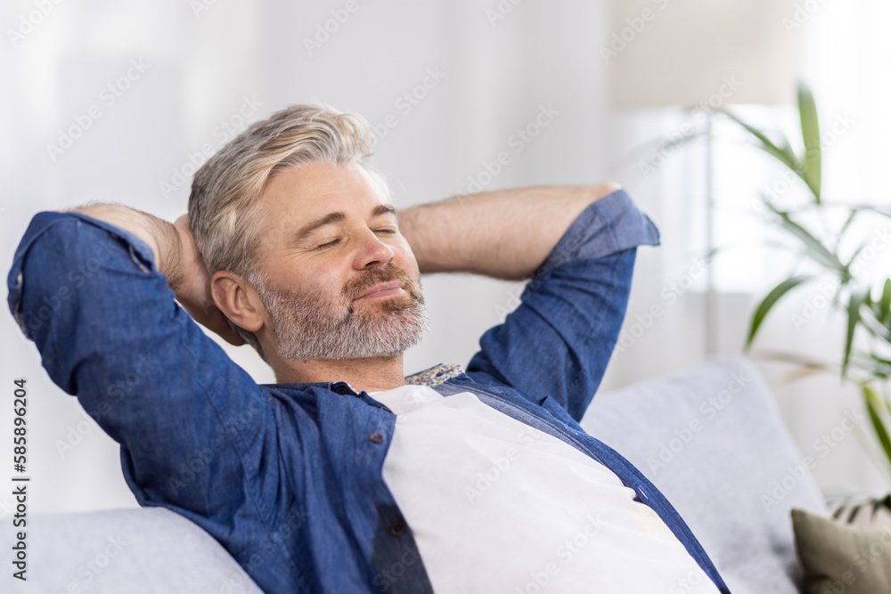 Close-up photo. Senior gray-haired man sits on the sofa at home and rests, takes a nap. He put his hands behind his head, closed his eyes, relaxed after a hard day's work. Close-up photo. Senior gray-haired man sits on the sofa at home and rests, takes a nap. He put his hands behind his head, closed his eyes, relaxed after a hard day's work.